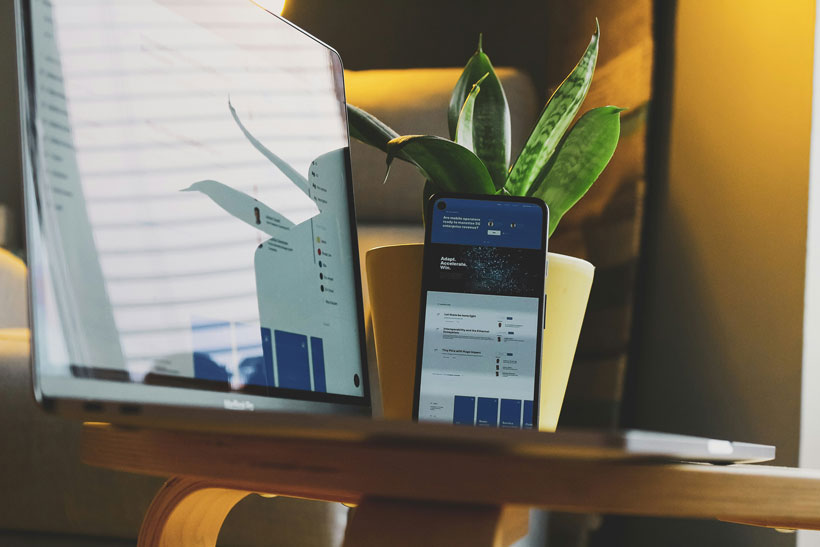 Écran d'ordinateur et smartphone affichant un tableau de bord de gestion administrative, posés sur un bureau en bois avec une plante en arrière-plan. Ambiance de travail numérique et connectée
