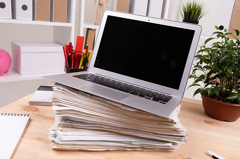 Ordinateur portable posé sur une pile de documents papier dans un bureau, illustrant la surcharge administrative avant la dématérialisation.