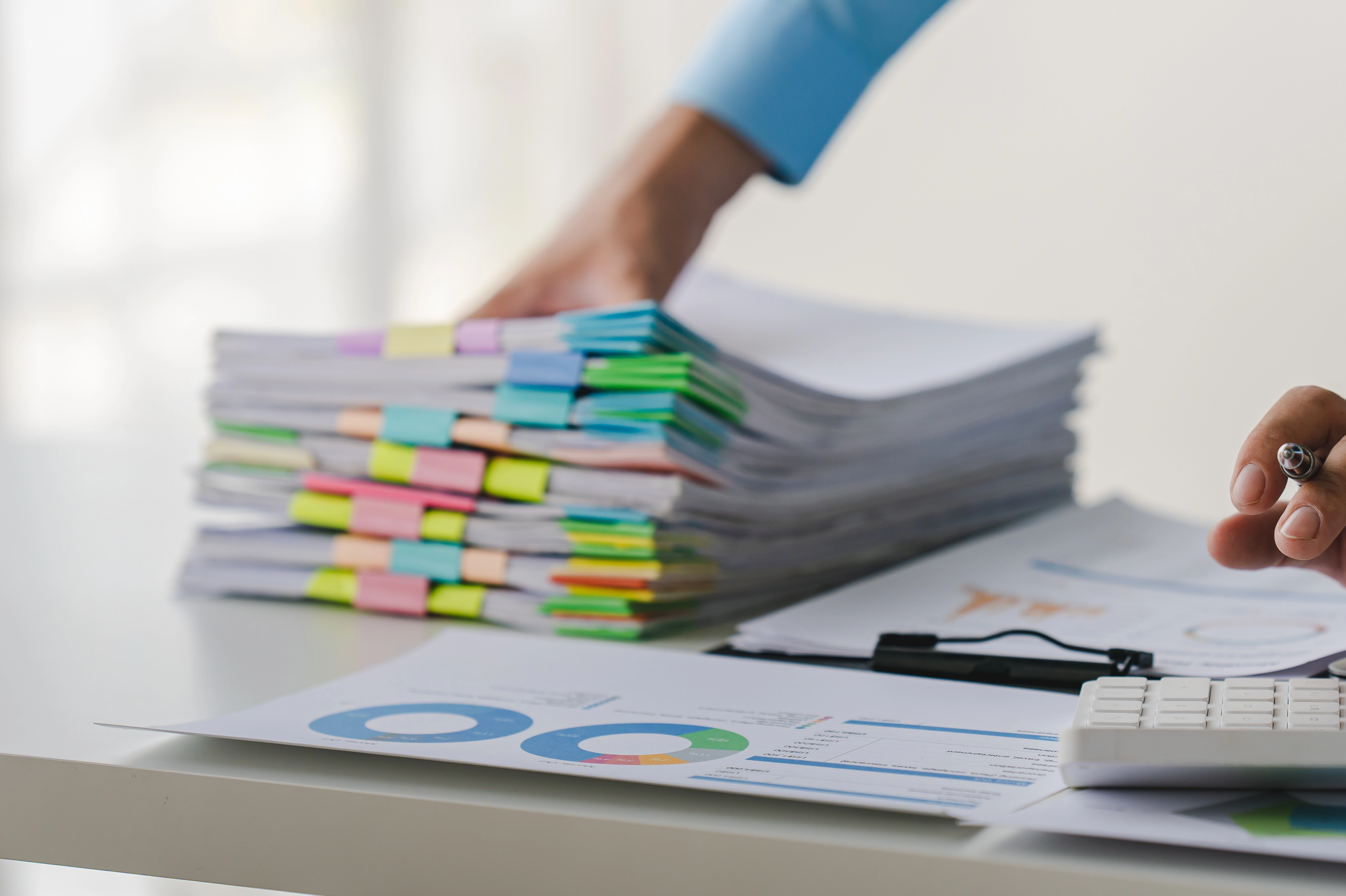 Main d’un professionnel manipulant une pile de dossiers classés par couleurs, sur un bureau d’entreprise avec documents et calculatrice